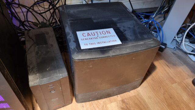 A cube-shaped black computer sits next to a battery pack on a laminated floor. A sign has been left atop it, reading "Caution: Generator connected to this installation."